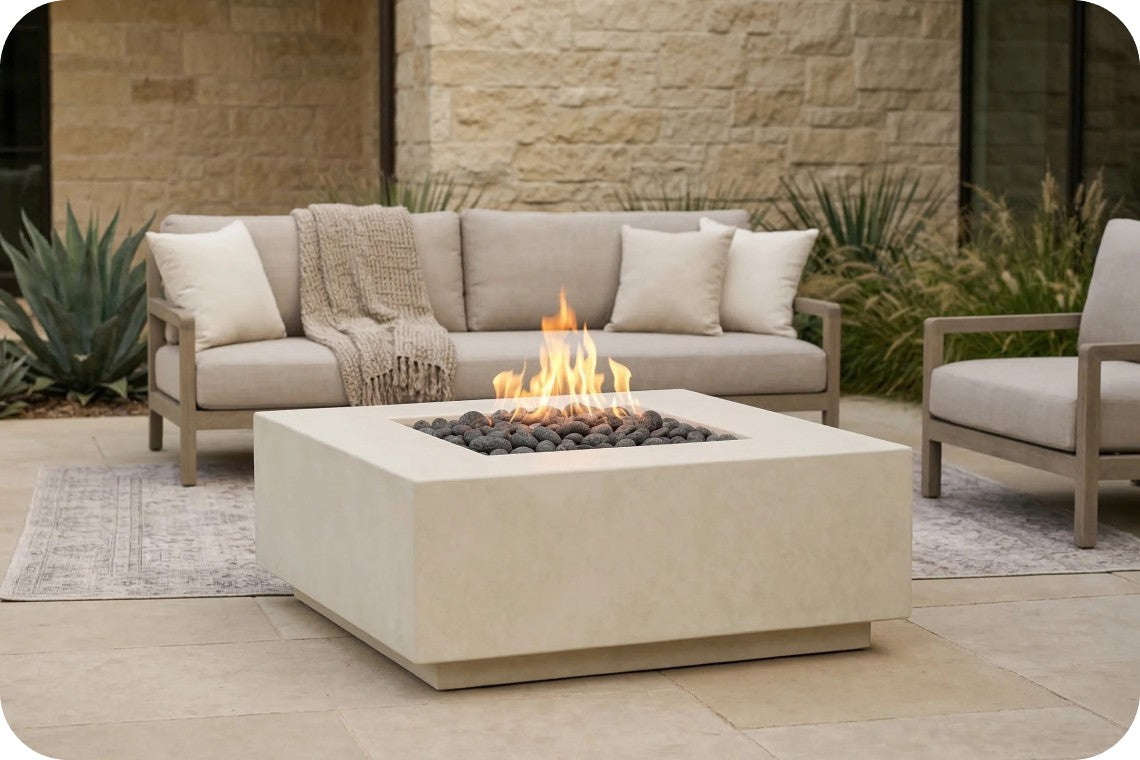 A modern outdoor patio scene featuring a square concrete fire pit table with a live flame and dark river rocks, positioned in front of a light beige sectional sofa and a matching armchair.