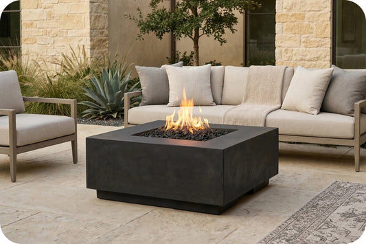 A square black fire pit table with an active flame, positioned in front of a light beige outdoor sofa and chair on a stone patio.