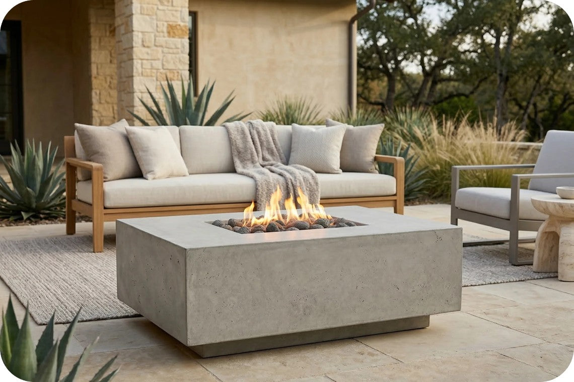 A modern outdoor patio scene featuring a square concrete fire pit table with a lit flame, positioned in front of a light wood and beige cushion outdoor sofa. The setting includes a neutral rug, potted agaves, and a matching outdoor armchair under warm natural light.