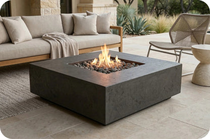 A square concrete fire pit table with a live flame and dark stones, set on a light stone patio next to a beige outdoor sofa and chair.