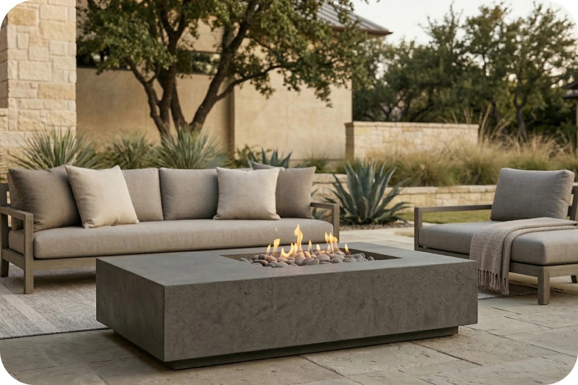Outdoor patio furniture set featuring a rectangular concrete fire pit table, a three-seat sofa with beige cushions, and a matching armchair with a throw blanket, arranged on a stone patio in a landscaped backyard setting.