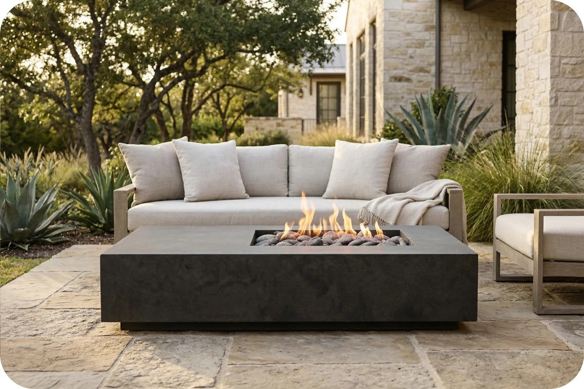 Outdoor patio furniture set featuring a modern rectangular black fire pit table with active flames, positioned in front of a light beige sectional sofa with matching throw pillows and a throw blanket. The scene is set on a stone patio with a stone house and green plants in the background.