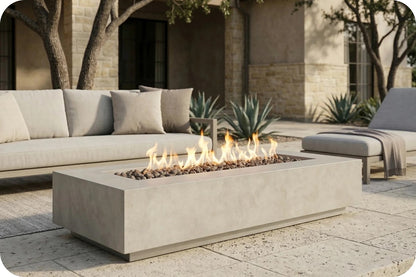 A rectangular concrete fire pit table with active flames, set on a stone patio in an outdoor patio setting with a sofa and lounge chair in the background.