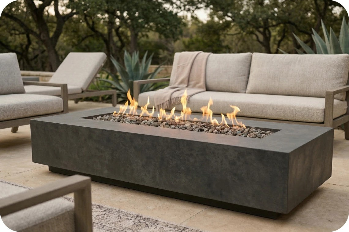 A rectangular concrete fire pit table with active flames and gray stones, placed in an outdoor patio setting with a light beige sofa and lounge chairs in the background.
