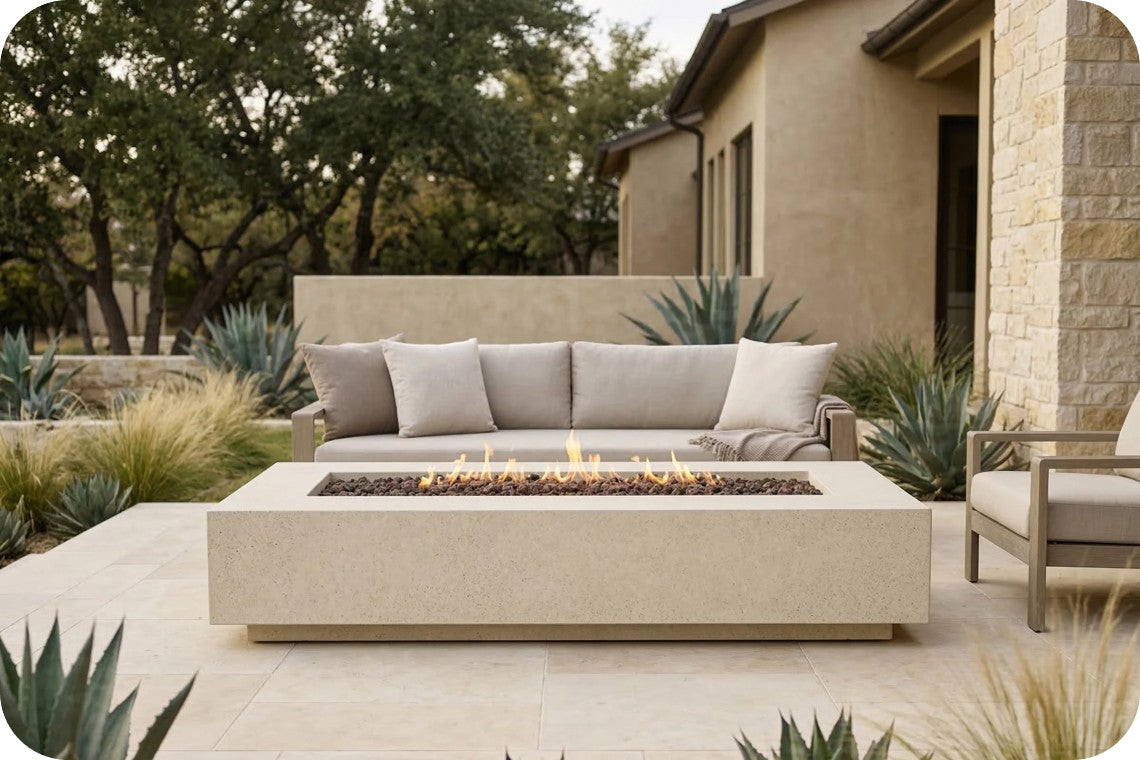 A modern outdoor patio scene featuring a rectangular concrete fire pit table with a lit fire, positioned in front of a matching outdoor sofa with beige cushions. The setting includes a stucco house, stone columns, and desert plants.