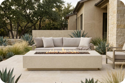 A modern outdoor patio scene featuring a rectangular concrete fire pit table with a lit fire, positioned in front of a matching outdoor sofa with beige cushions. The setting includes a stucco house, stone columns, and desert plants.