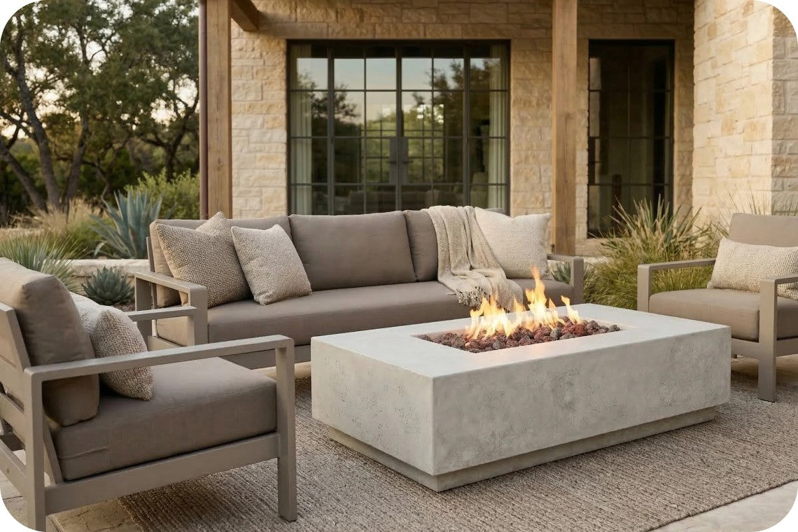 Outdoor patio furniture set with a concrete fire pit table, a gray sectional sofa, and two matching armchairs in front of a stone house.