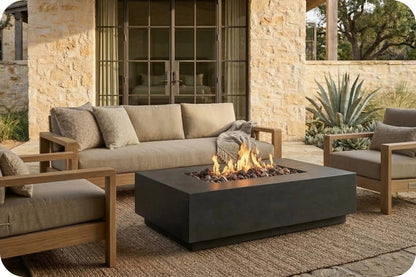 Outdoor patio furniture set including a beige cushioned sofa, two matching armchairs with wooden frames, and a rectangular black fire pit table with visible flames, arranged on a woven rug in front of a stone building.