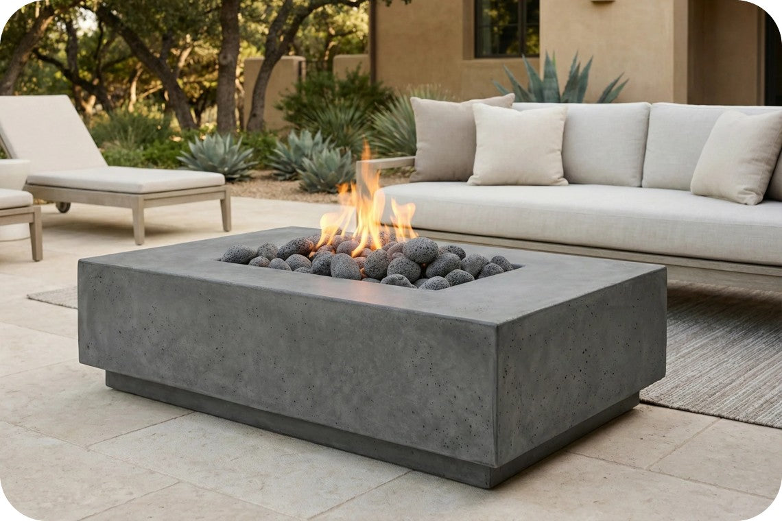 A rectangular concrete fire pit table with a square base, filled with dark river rocks and a visible flame, set on a patio in front of a light beige outdoor sofa.