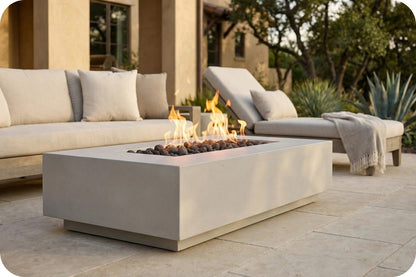 A rectangular concrete outdoor fire pit table with active flames and dark river rocks, set on a stone patio in front of a light beige sectional sofa and chaise lounge.