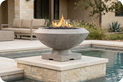 A round concrete fire pit bowl with a pedestal base, filled with black stones and active orange flames, situated on a stone pool deck next to a swimming pool in an outdoor patio setting.