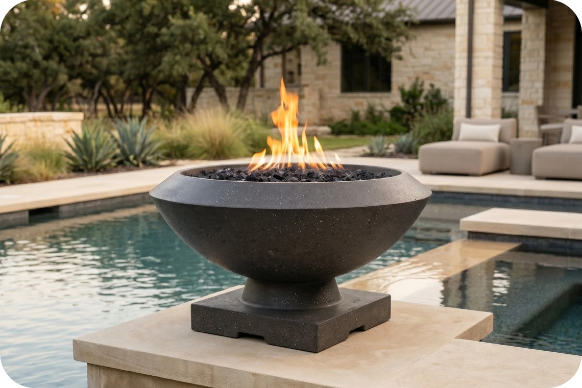 A dark gray stone bowl fire pit with a square pedestal base, filled with black stones and a bright orange flame, situated on a stone deck next to a swimming pool in an outdoor patio setting.