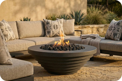 A round concrete fire pit with active flames, surrounded by black river rocks, set in a patio outdoor seating area with beige sectional cushions and neutral throw pillows.